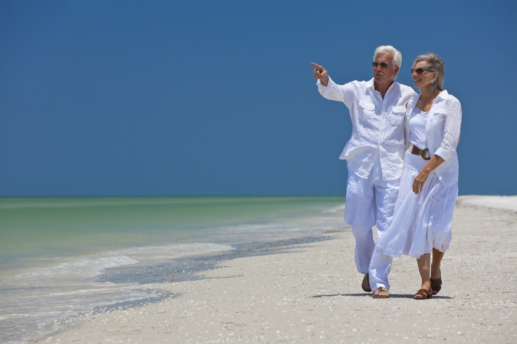 Happy senior man and woman couple walking together looking out to sea on a deserted tropical beach with bright clear blue sky, the man is pointing to the horizon